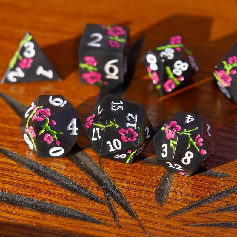 May include: A set of black polyhedral dice with white numbers and pink floral designs. The dice are arranged on a wooden surface, showcasing various shapes, including a D4, D6, D8, D10, D12, and D20.