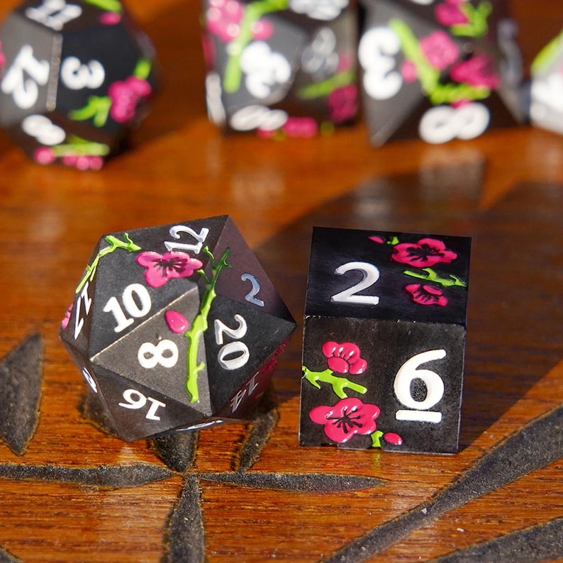 May include: Black polyhedral dice set with white numbering and pink floral accents. The set includes a D20 and a D6 die. The dice are decorated with pink flowers and green vines. The dice are on a wooden surface.