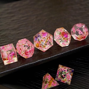 Könnte beinhalten: Ein Set aus transparenten Harzwürfeln mit rosa Zahlen und eingebetteten getrockneten Blumen. Die Würfel haben verschiedene geometrische Formen, darunter ein Würfel, ein d20 und eine Pyramide. Die Blumen sind in Rosa-, Gelb- und Lilatönen gehalten.