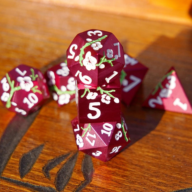 May include: A collection of burgundy polyhedral dice with white floral designs and white numbering. The dice are arranged on a wooden surface, with some dice stacked and others scattered. The dice are for tabletop games.