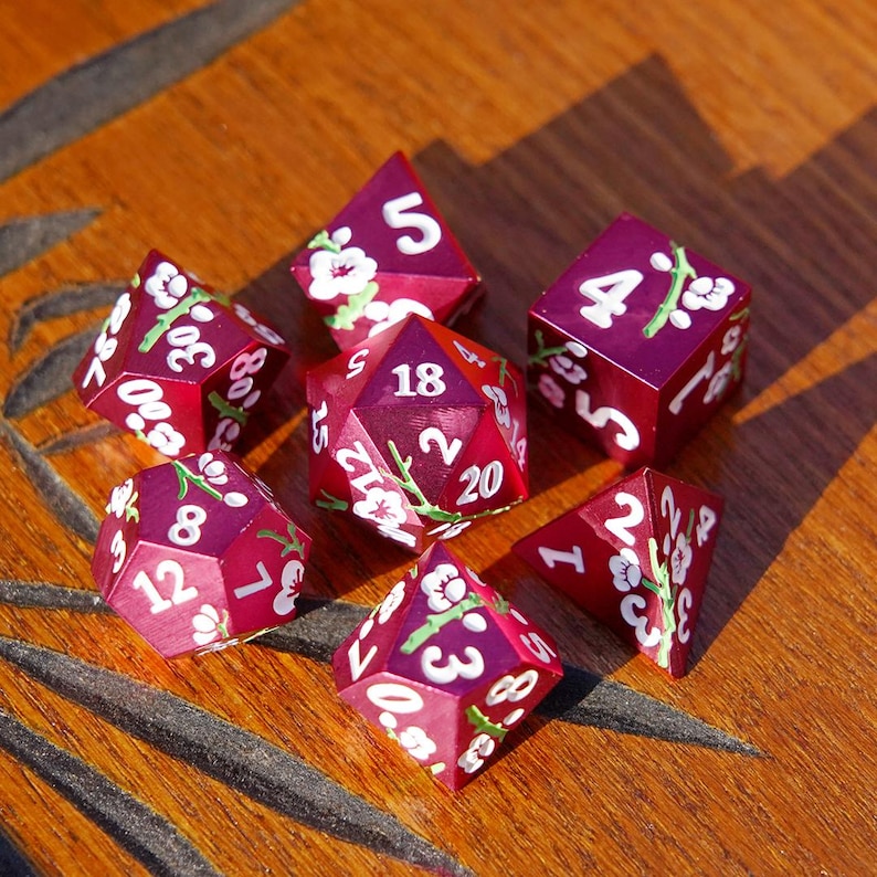 May include: A set of seven burgundy dice with white numbers and floral accents. The dice are arranged on a wooden surface, showcasing their various shapes and sizes, including a D20, D6, and D4.