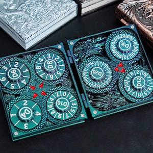 May include: A teal-colored, hinged gaming dice case featuring six circular, rotating dials labeled D4, D6, D8, D10, D12, D20, and D%. The case has a textured, dragon-themed surface with small red triangular accents and magnetic closures.