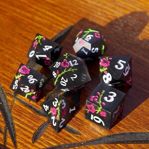 May include: Set of seven black polyhedral dice with white numbering and pink floral designs. The dice are for role-playing games and include a D4, D6, D8, D10, D12, D20, and a percentile die.