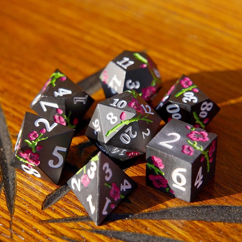 May include: A set of seven black polyhedral dice with white numbering and pink floral designs. The dice are arranged on a wooden surface, with various faces visible, including a D20, D12, D10, D8, D6, D4, and D100.