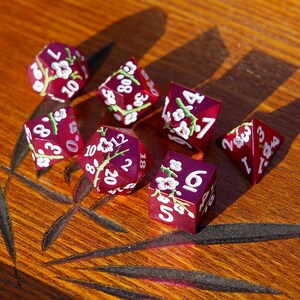May include: A set of seven burgundy dice with white numbers and floral designs. The dice are in various polyhedral shapes, including a d20, d10, d8, d6, and d4. The dice are arranged on a wooden surface.