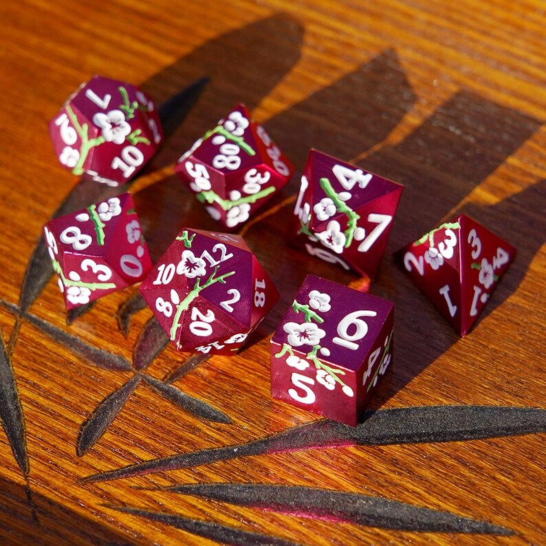 May include: A set of seven burgundy dice with white numbers and floral designs. The dice are in various polyhedral shapes, including a d20, d10, d8, d6, and d4. The dice are arranged on a wooden surface.