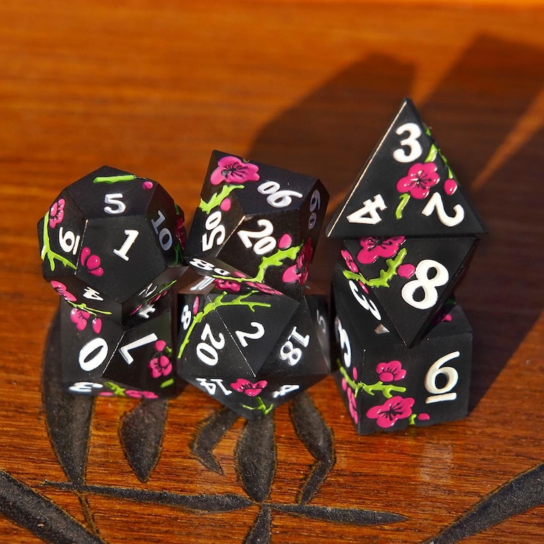 May include: A set of black polyhedral dice with white numbering and pink floral designs. The dice are arranged on a wooden surface, showcasing their various shapes and sizes, including a d20, d10, and d4.