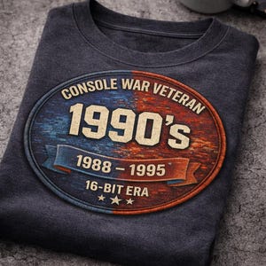 May include: Dark gray t-shirt with a retro design. The design features an oval with the text "CONSOLE WAR VETERAN 1990'S 1988-1995 16-BIT ERA" in a distressed style. The shirt is folded on a textured surface.