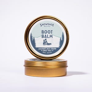 May include: Two gold-colored tins of Snowshoe Brand Boot Balm. The top tin displays a label with the words "Boot Balm" and a graphic of a boot. The label also includes the text "Leather's Best Friend Since 1927" and "Bend, Oregon."