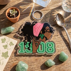May include: A clear acrylic keychain featuring a stylized illustration of a person with dark curly hair wearing a pink beanie and a green ivy leaf earring. The design includes a hand making a gesture and the green numbers 1908 with a white outline.