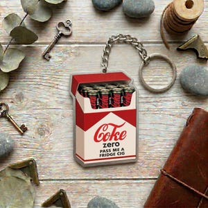 May include: A keychain featuring a rectangular charm shaped like a cigarette pack, colored red, white, and cream. The charm displays the text "Coke zero PASS ME A FRIDGE CIG" and depicts several black soda cans inside the open top of the pack.