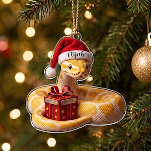 May include: A flat acrylic Christmas ornament featuring a yellow and white patterned snake wearing a red Santa hat with the name Elijah printed on the brim. The snake is coiled around a red gift box with the text Sneaky Santa printed on the front.