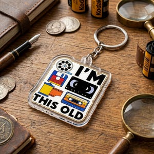 May include: A clear acrylic keychain featuring a graphic design with a film reel, floppy disk, camera film roll, VHS tape, and audio cassette. The text "I'M THIS OLD" is printed in bold black letters on the front of the square-shaped keychain.
