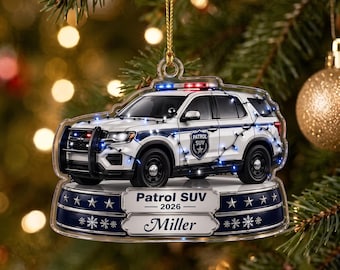 May include: A clear acrylic Christmas ornament featuring a white police patrol SUV with a black push bumper, wrapped in blue string lights. The vehicle sits on a dark blue base with white stars and snowflakes, labeled Patrol SUV 2026 and Miller.