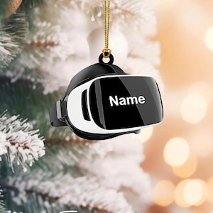 May include: A black and white VR headset Christmas ornament with the word "Name" printed on the screen. The ornament hangs from a gold chain. The background is blurred with Christmas tree branches and lights.