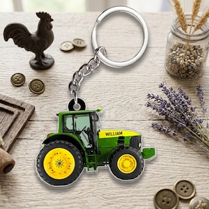 May include: A silver metal keychain featuring a flat, acrylic tractor charm. The tractor is green with yellow wheels and a yellow hood section displaying the name WILLIAM in black text. The charm is attached to a standard circular split ring.