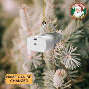May include: White and gray VR headset Christmas ornament hanging from a gold string. The ornament has a gray strap and the text "Name" on the front. A "Santa Approved" sticker is in the background. The text "Name Can Be Changed" is on a yellow label.