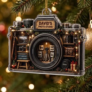 May include: A camera-shaped Christmas ornament featuring a wooden plaque labeled DAVID'S PHOTO STUDIO. The interior displays a miniature photography studio with a camera on a tripod, framed photos, books, a camera bag, and a gift box, all accented with tiny lights.