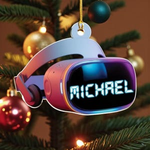 May include: A VR headset Christmas ornament with the name "MICHAEL" displayed on the screen. The ornament is primarily pink, purple, and blue, with a gold string for hanging. It is set against a backdrop of Christmas tree branches and ornaments.