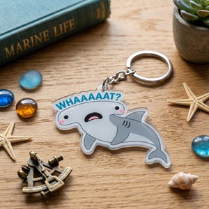 May include: A clear acrylic keychain featuring a cartoon hammerhead shark with a surprised expression and the text WAAAAAT? in blue. The shark is light gray with a white belly, pink cheeks, and a black mouth. It includes a silver metal ring.
