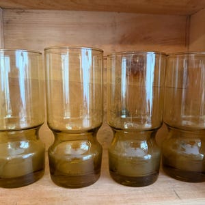 May include: A set of four tall, cylindrical drinking glasses made of translucent amber-coloured glass. Each glass features a distinct, smooth indentation around the lower section, creating a tapered base that widens towards the top rim.