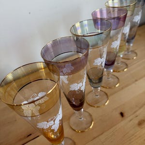 May include: A set of six tall, slender champagne flutes arranged in a row. Each glass features a delicate white etched grape leaf pattern, a thin gold rim, and a distinct iridescent or coloured tint, including amber, purple, and blue hues.