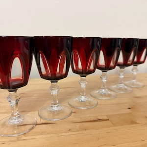 May include: A set of six wine glasses featuring deep red bowls with clear glass cut-out arch patterns. Each glass has a clear, faceted stem and a circular base. The glasses are arranged in a diagonal line, showcasing the contrast between the dark red glass and the transparent clear glass elements.
