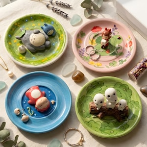 Puede incluir: Cuatro platos decorativos de cerámica con diseños caprichosos. Uno presenta una figura gris de Totoro, otro una niña sobre una hoja de nenúfar, un tercero con dos figuras y el último con tres espíritus del bosque blancos sobre un tronco. Cada plato es de un color diferente.