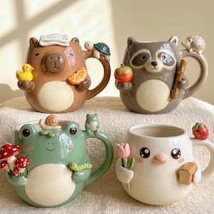 May include: Four ceramic mugs with animal designs. A brown bear mug holds a yellow duck and orange slice. A raccoon mug holds an apple and stick. A green frog mug has a snail and mushroom. A white bird mug holds flowers and bread.