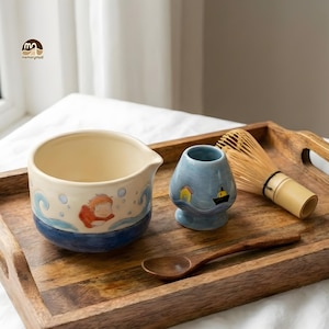 May include: A ceramic matcha bowl with a painted orange character and blue waves, a light blue whisk holder with a small house and boat illustration, a wooden spoon, and a bamboo whisk on a rectangular wooden tray with cutout handles.