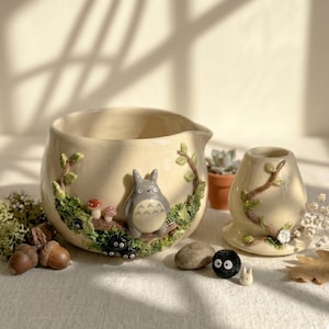 Pode incluir: Conjunto de vasos e floreiras de cerâmica creme, decorado com uma cena de floresta com uma figura cinzenta de Totoro, cogumelos e outros personagens. A floreira tem um bico e o vaso é cilíndrico. O conjunto está sobre uma superfície branca.