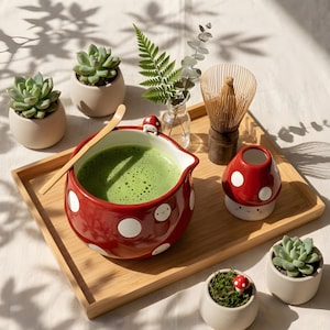 Op de afbeelding: Een rode keramische matcha-kom met witte stippen en een paddenstoelvormige handgreep, gevuld met groene matcha-thee. Een houten garde en een kleine paddenstoelvormige container staan ook op de bamboe lade, samen met kleine vetplanten in pot.