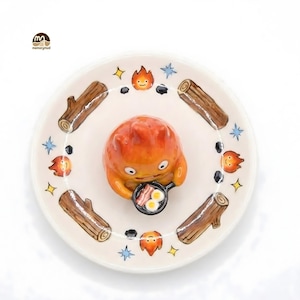 May include: A ceramic plate featuring a cartoon character holding a frying pan with bacon and eggs. The plate is decorated with logs, flames, stars, and black stones. The character is orange and the plate is white.