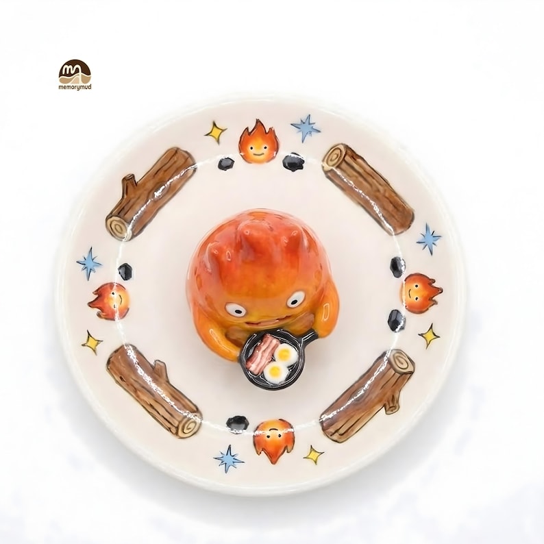 May include: A ceramic plate featuring a cartoon character holding a frying pan with bacon and eggs. The plate is decorated with logs, flames, stars, and black stones. The character is orange and the plate is white.