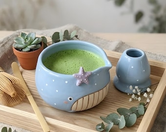May include: A light blue ceramic whale-shaped matcha bowl with white polka dots, a small purple starfish accent, and a beige ribbed belly. Beside it sits a matching light blue whisk holder, a bamboo whisk, and a small potted succulent plant.