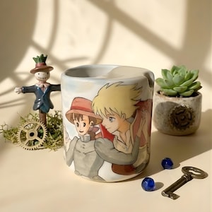 May include: A ceramic mug featuring a colorful illustration of two anime characters. The mug is white with a curved handle and a wide opening. A small succulent plant in a decorative pot, a scarecrow figurine, and a vintage key are also in the image.