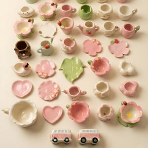 May include: A collection of whimsical ceramic miniatures in various shapes and colors. Includes flower-shaped bowls, teacups, saucers, and animal figurines. The color palette features soft pinks, greens, and whites, with some pieces adorned with small details like strawberries and ladybugs.