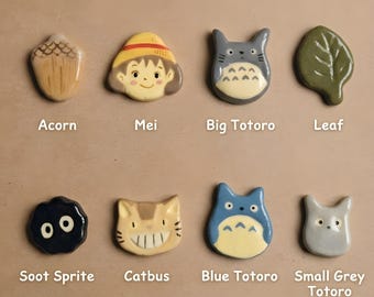 May include: A set of eight ceramic charms featuring various characters and objects including an acorn, Mei, Big Totoro, a leaf, a soot sprite, Catbus, Blue Totoro, and Small Grey Totoro, each labeled with its name in white text below the item.