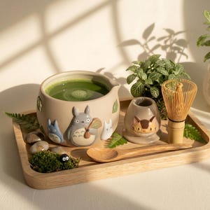 Op de afbeelding: Een matcha-theeset met een beige keramische kom gevuld met groene matcha. De kom is versierd met stripfiguren. De set bevat een garde, een keramische houder, een houten lepel en een houten dienblad. Kleine planten en stenen omringen de set.