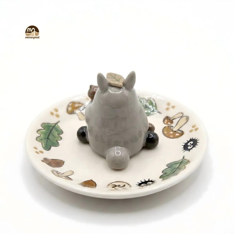 May include: A gray ceramic Totoro figurine with a leaf on its head sits on a small, off-white plate. The plate is decorated with painted leaves, mushrooms, and other whimsical elements. The logo "memorymud" is in the upper left corner.