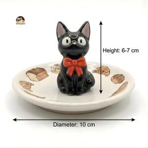May include: A black ceramic cat figurine with a red bow tie sits on a small, off-white dish. The dish has a diameter of 10 cm and features bread and pastry illustrations. The cat figurine is 6-7 cm tall.