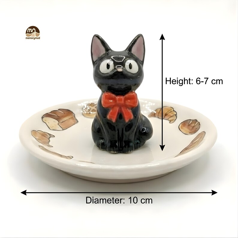 May include: A black ceramic cat figurine with a red bow tie sits on a small, off-white dish. The dish has a diameter of 10 cm and features bread and pastry illustrations. The cat figurine is 6-7 cm tall.