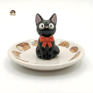 May include: A black cat figurine with a red bow tie sits atop a small, off-white ceramic dish. The dish is decorated with illustrations of bread and pastries. The cat has large, round eyes and pointed ears.