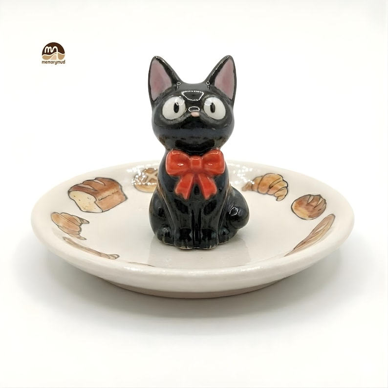 May include: A black cat figurine with a red bow tie sits atop a small, off-white ceramic dish. The dish is decorated with illustrations of bread and pastries. The cat has large, round eyes and pointed ears.