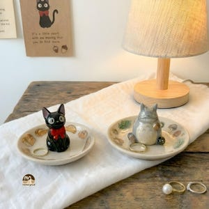 May include: Two decorative ceramic ring dishes. One features a black cat with a red bow, the other a gray cartoon character. Both dishes are off-white with decorative elements. A lamp and rings are also visible.