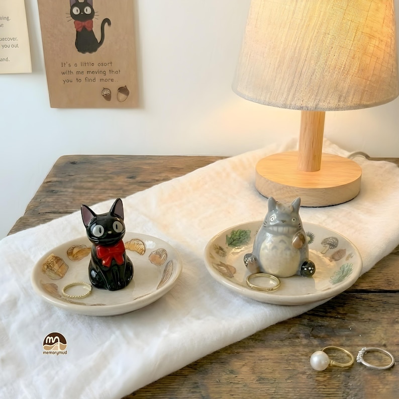 May include: Two decorative ceramic ring dishes. One features a black cat with a red bow, the other a gray cartoon character. Both dishes are off-white with decorative elements. A lamp and rings are also visible.