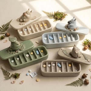 May include: Four rectangular ceramic pill organisers with lids featuring sculpted forest creatures. Each container has seven compartments labelled M, T, W, T, F, S, S, containing various small round and oval pills in white, blue, and yellow colours.