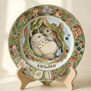 Puede incluir: Un plato decorativo de cerámica con una ilustración caprichosa de un personaje Totoro durmiendo en una escena de bosque. El plato tiene un borde verde con setas y otros elementos forestales. El plato se exhibe en un soporte de madera.