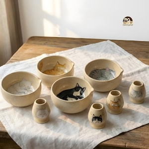 Puede incluir: Colección de cuencos y pequeños jarrones de cerámica, cada uno con un diseño de gato diferente. Los cuencos tienen un exterior beige y una forma redondeada, con ilustraciones de gatos en el interior. Los jarrones son más pequeños y tienen un diseño similar. Los gatos están representados en varias poses y colores.