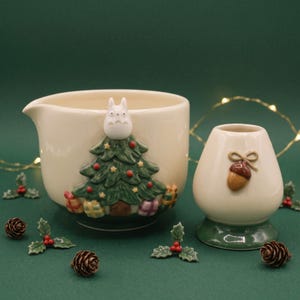 May include: A cream-colored ceramic pouring bowl featuring a raised Christmas tree with a white character topper and colorful gift boxes, paired with a matching cream-colored whisk holder decorated with a brown acorn and a bow.